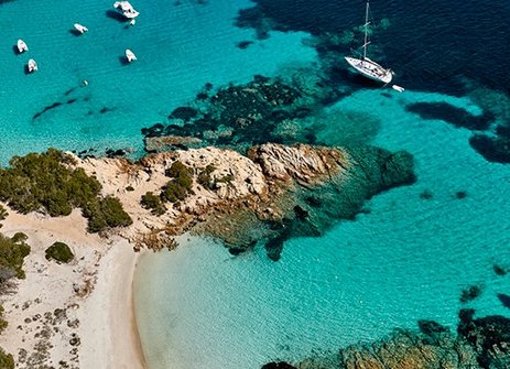 Strand in Sardinien mit Booten im Wasser und einer felsigen Küste im Hintergrund.