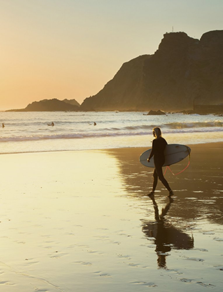 Eine Person geht am Strand der Algarve mit einem Surfbrett in der Hand entlang.