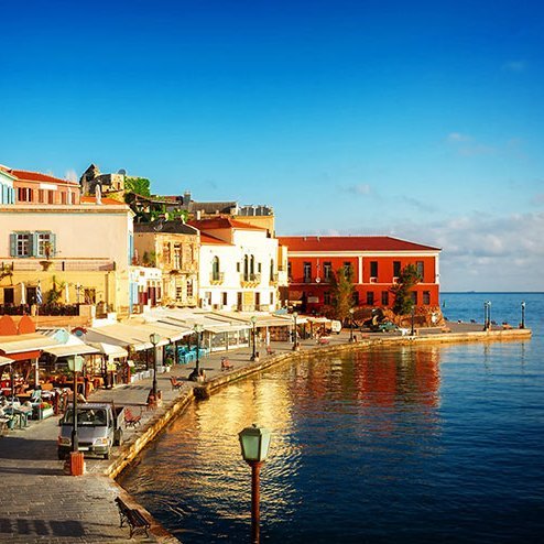 Blick auf den Hafen und die Gebäude einer Stadt in Kreta, umgeben von klarem Wasser und strahlendem Himmel.