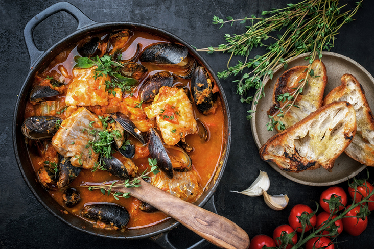 Herzhaftes Fisch- und Muscheleintopf in einer Pfanne, serviert mit Brot und saftigen Tomaten.