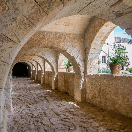 Langer Tunnel im Kloster Arkadi mit Bögen und Töpfen voller Pflanzen, die eine grüne Atmosphäre schaffen.