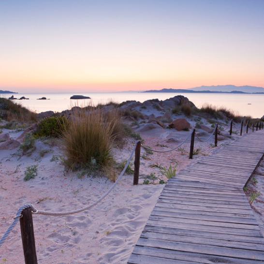 Holzsteg, der bei Sonnenuntergang zum Strand von La Maddalena führt, umgeben von sanften Wellen und warmen Farben.