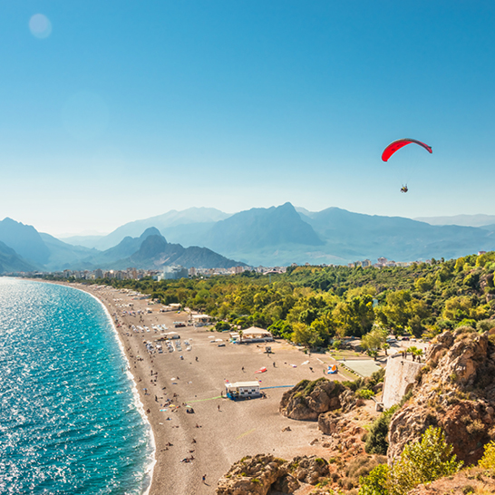 Ein Paraglider schwebt über einem Strand mit einer Bergkette im Hintergrund, Badeferien in Antalya.