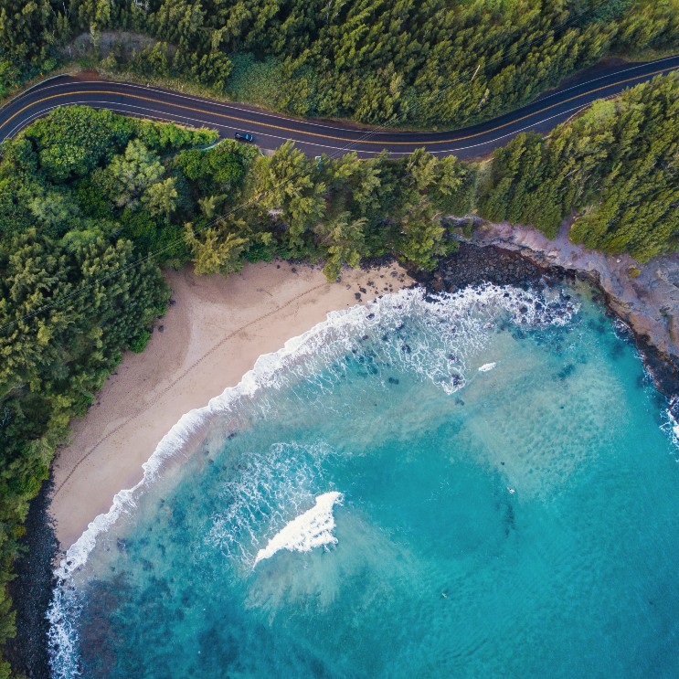 Luftperspektive über einer blauen Bucht in Hawaii mit einer gewunden Strasse darüber und grünem Wald